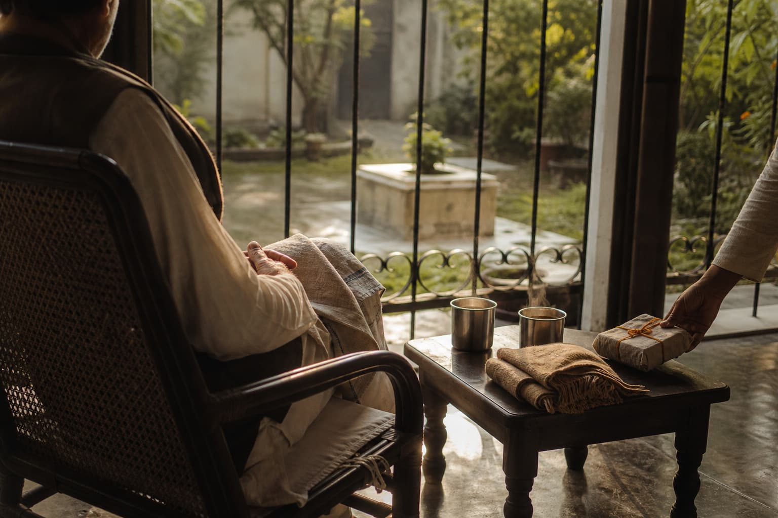 An elderly man in a kurta and waistcoat sits in a cane chair on a verandah, seen from behind, as a younger person's hand sets a gift in handmade paper, tied with orange thread, on a table beside two steel tumblers of chai and a folded shawl.